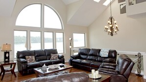 A spacious living room with two black leather sofas, a glass coffee table, and a chandelier. Large windows offer a view of a lake. - Luxurious 5 bedroom, 5+ baths, gourmet kitchen, great location (Camdenton)
