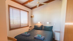 A bedroom with a bed, two bedside tables, a nightstand, a ceiling fan, a window with blinds, and a wall-mounted light.