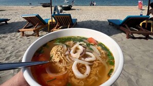 En skål suppe med nudler og fisk og skaldyr på en strand med parasoller og liggestole.