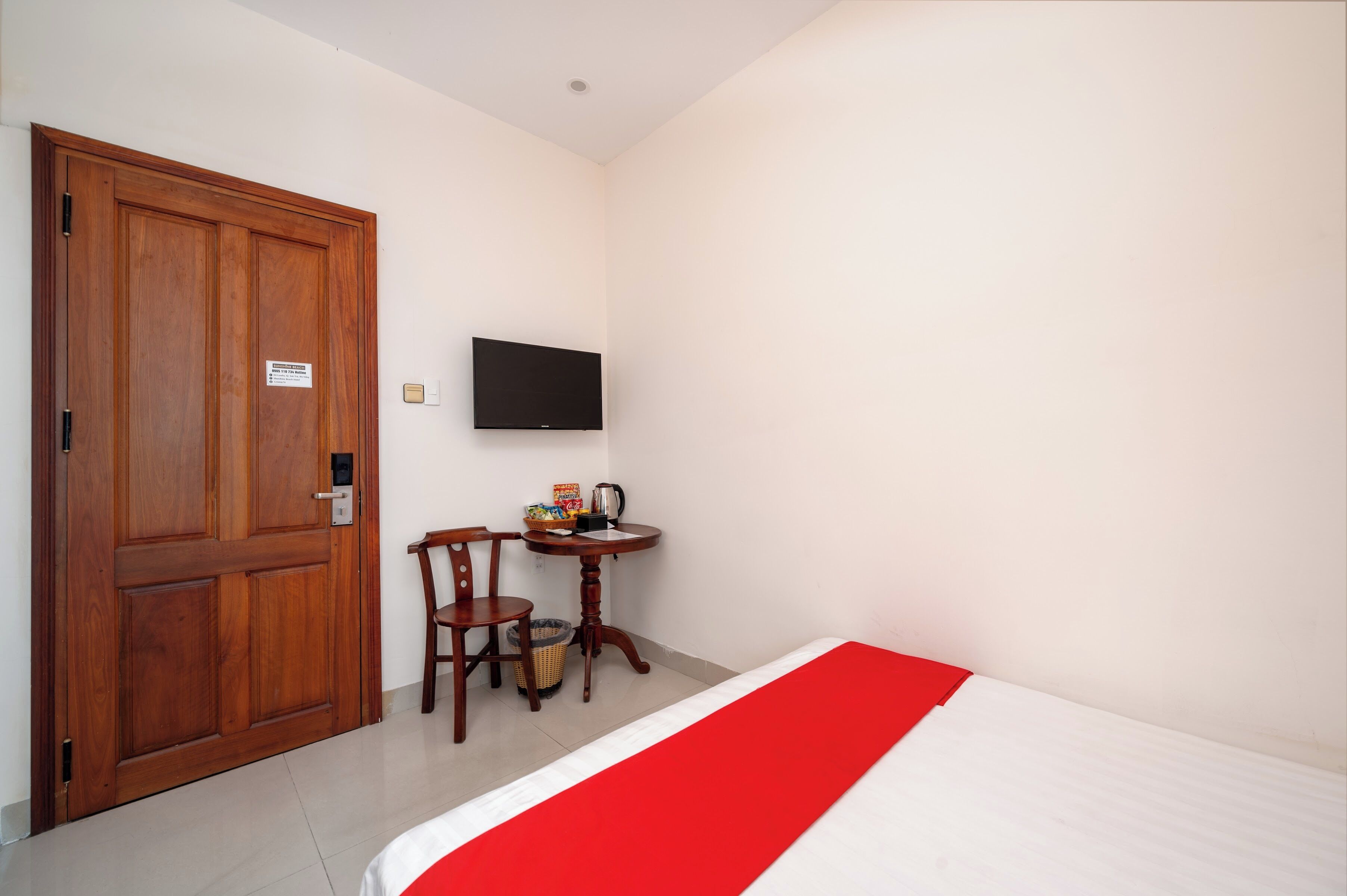 A hotel room with a wooden door, a television mounted on the wall, a small round table with a chair, and a bed with a red and white bedspread.