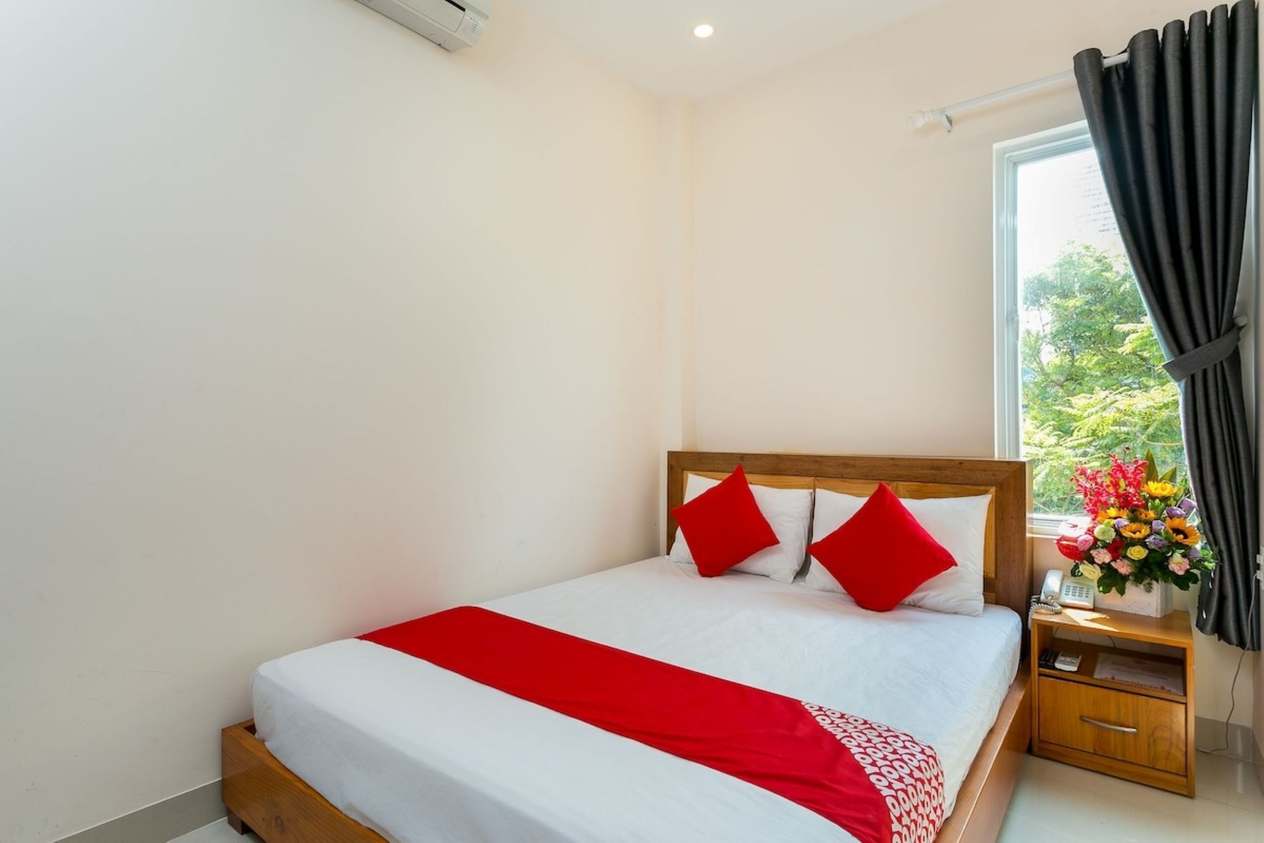 A neatly made bed with a red and white bedspread, a wooden headboard, and a nightstand with a telephone and a vase of flowers.