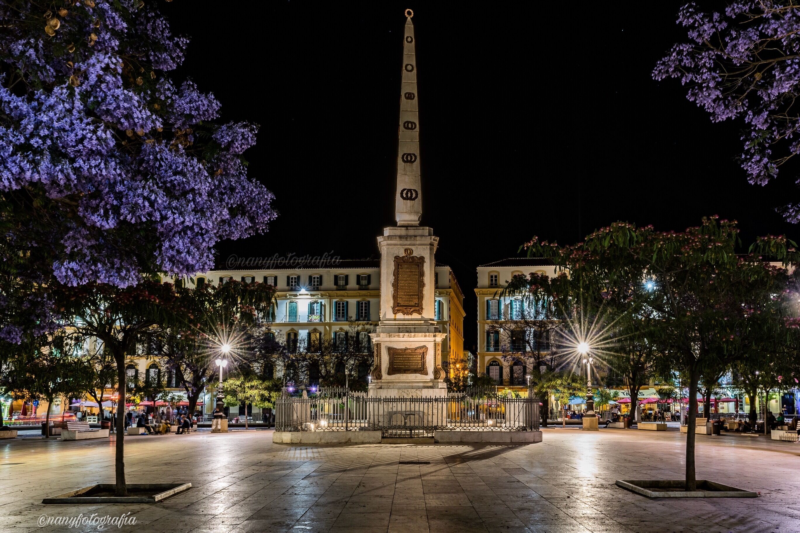 A square with a central monument, surrounded by trees and buildings with illuminated facades.
