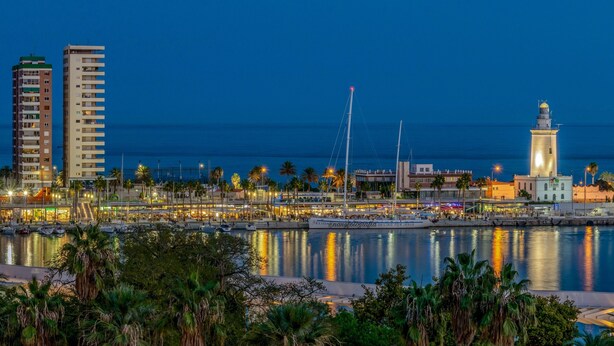 A coastal cityscape at night with a prominent lighthouse, a marina with boats, and modern buildings along the waterfront. - Charming apartment in the Historic Center ideal for couples and families (Málaga)