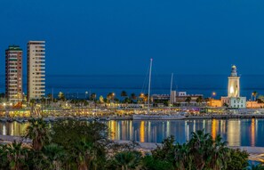 A coastal cityscape at night with a prominent lighthouse, a marina with boats, and modern buildings along the waterfront.