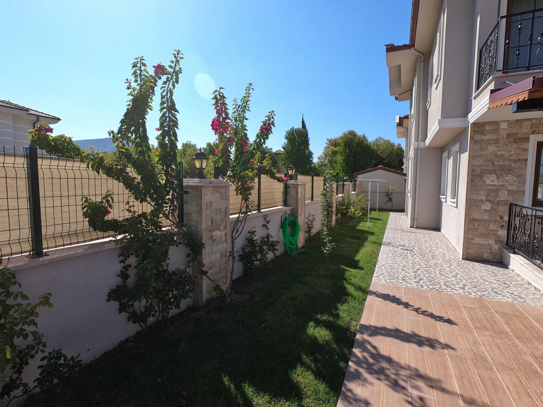 A residential area with a paved walkway, a stone wall, and a small garden with flowering plants.