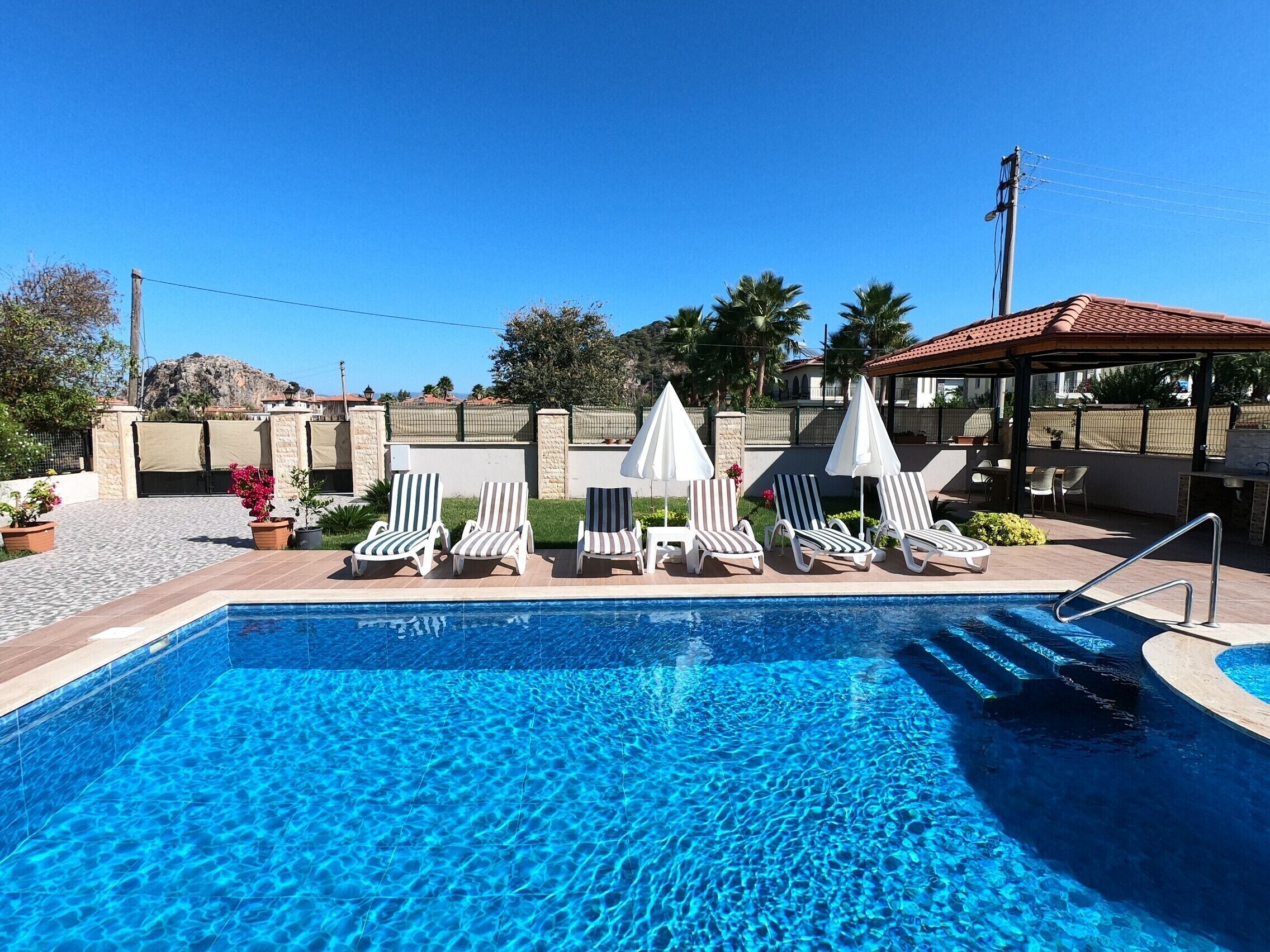 A swimming pool with lounge chairs and umbrellas, surrounded by a stone wall and a covered seating area.