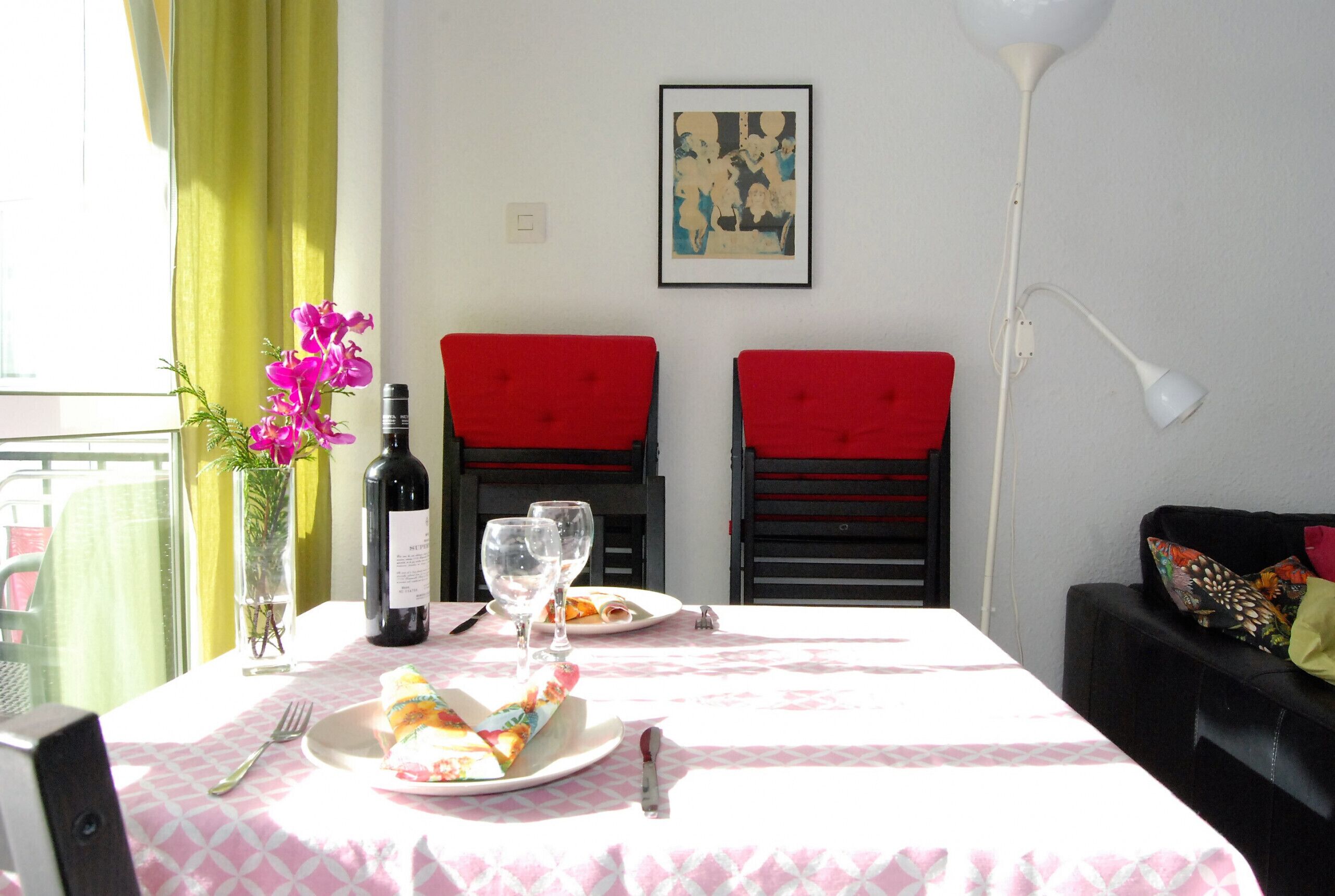 Una mesa de comedor con mantel a cuadros rosa y blanco, dos sillas tapizadas de rojo, un jarrón con flores rosas, una botella de vino y dos copas de vino.