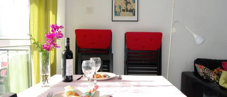 Una mesa de comedor con mantel a cuadros rosa y blanco, dos sillas tapizadas de rojo, un jarrón con flores rosas, una botella de vino y dos copas de vino.