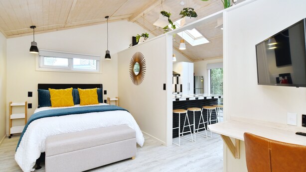 A modern bedroom with a bed, bedside tables, a bench, a TV, and a kitchenette area with stools. - Aspen Studio (Christchurch)