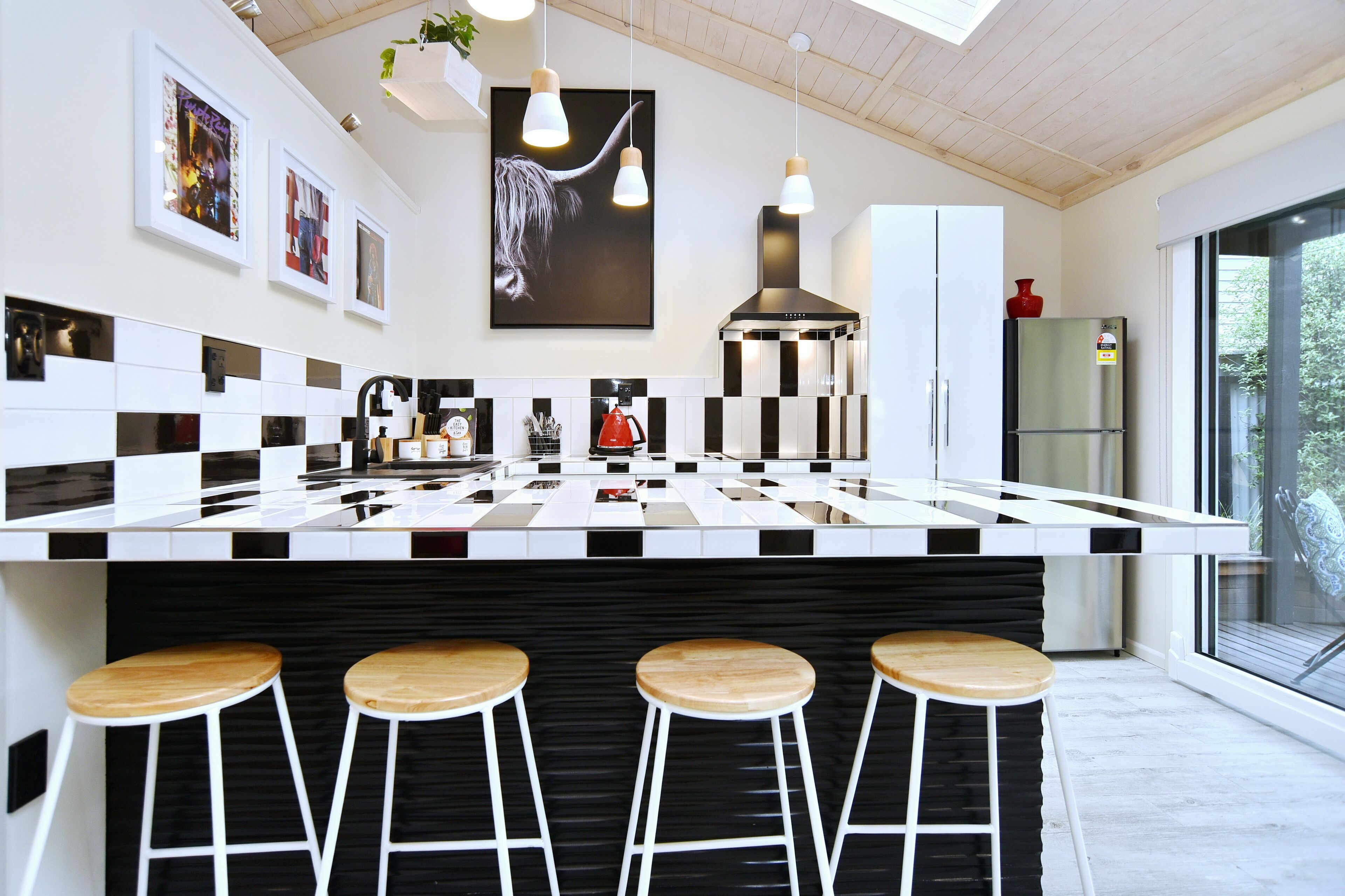 A modern kitchen with a black and white checkered countertop, four wooden stools, pendant lights, and a large window.