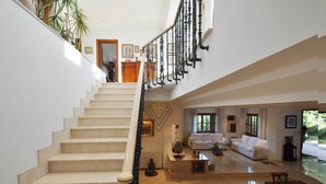 A spacious living area with a high ceiling, a staircase with ornate black railings, and a view of the outdoors through large windows. - Can Gual de Inca, Elegant Vila, Mallorca (Inca)