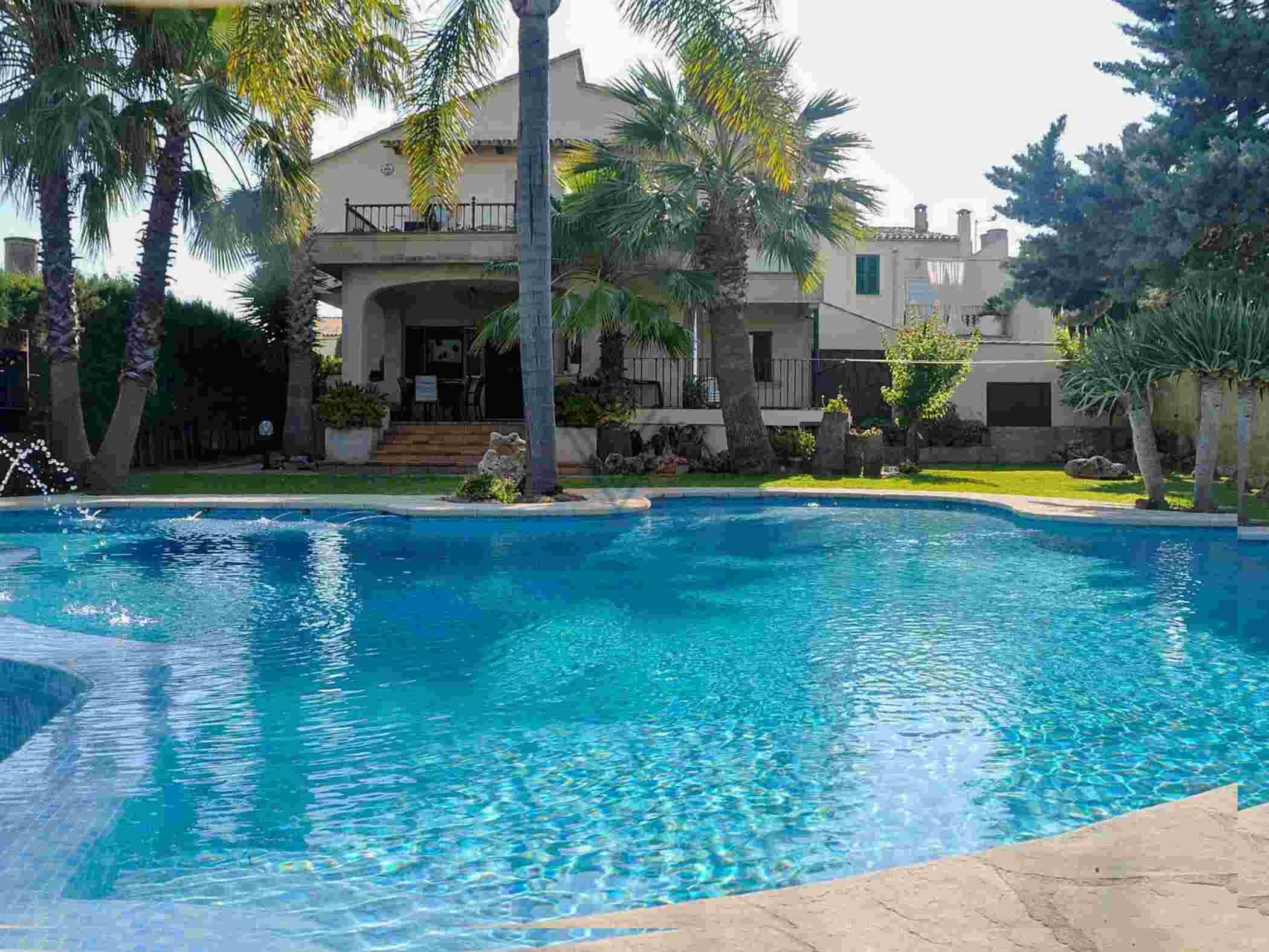 A swimming pool with a clear blue water surface, surrounded by palm trees and a large building with a balcony in the background.