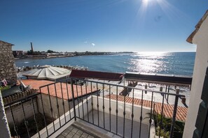 A balcony with a view of the sea, a stone building, and a red-tiled roof.