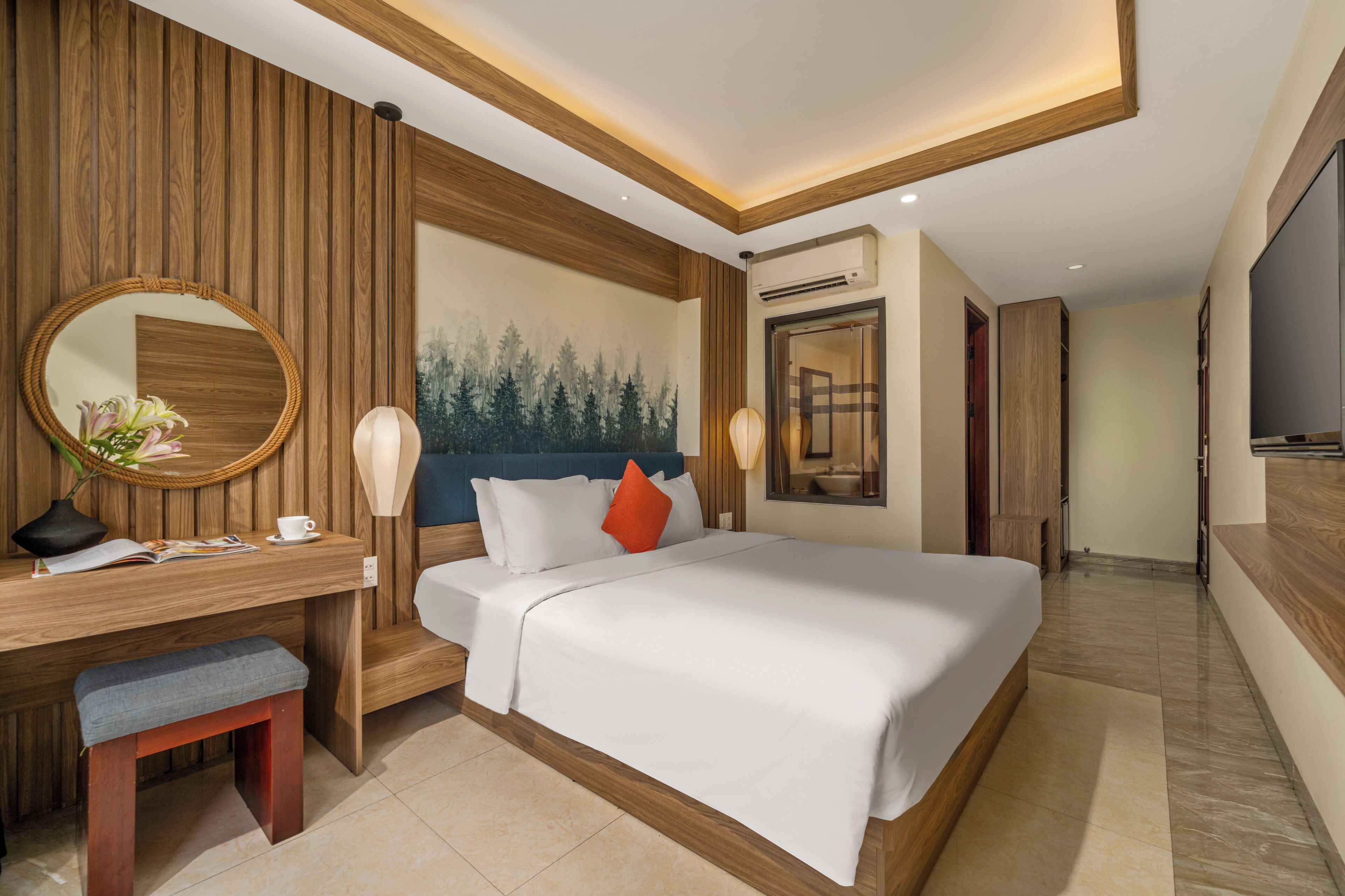A hotel room with a bed, a desk, a chair, a mirror, a painting, and an air conditioning unit.