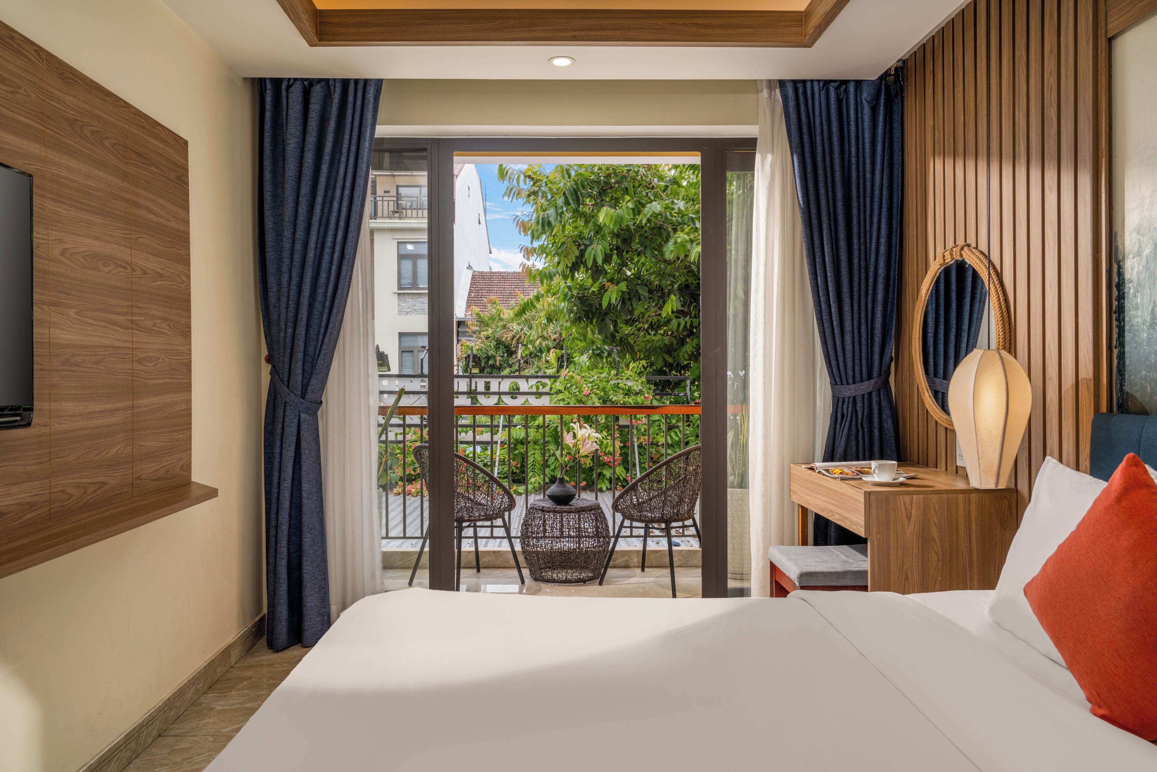A hotel room with a bed, a wooden headboard, a small table with a lamp, a chair, and a balcony with a view of greenery and buildings.