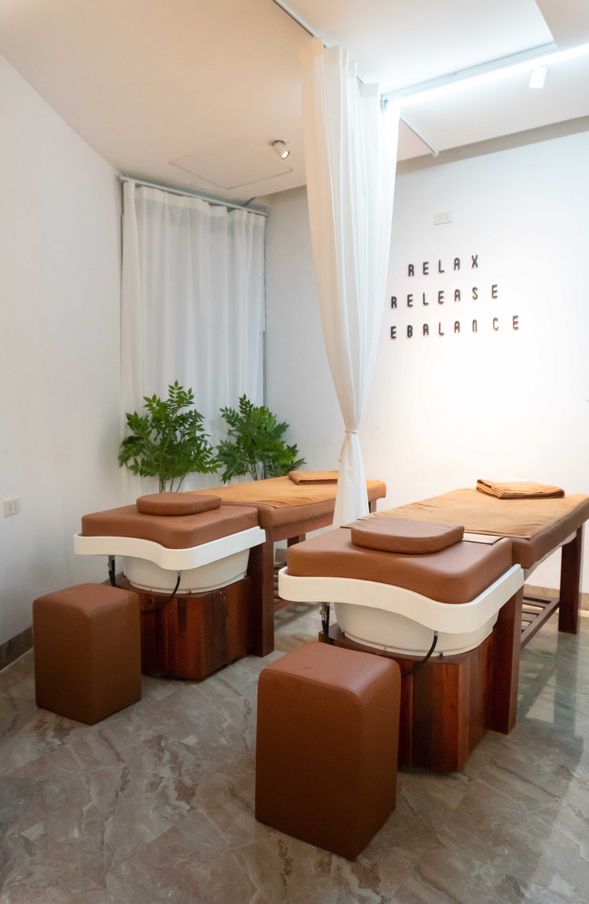 A massage therapy room with wooden tables and brown cushions, a white curtain, and a wall with the words "RELAX RELEASE EBALANCE".