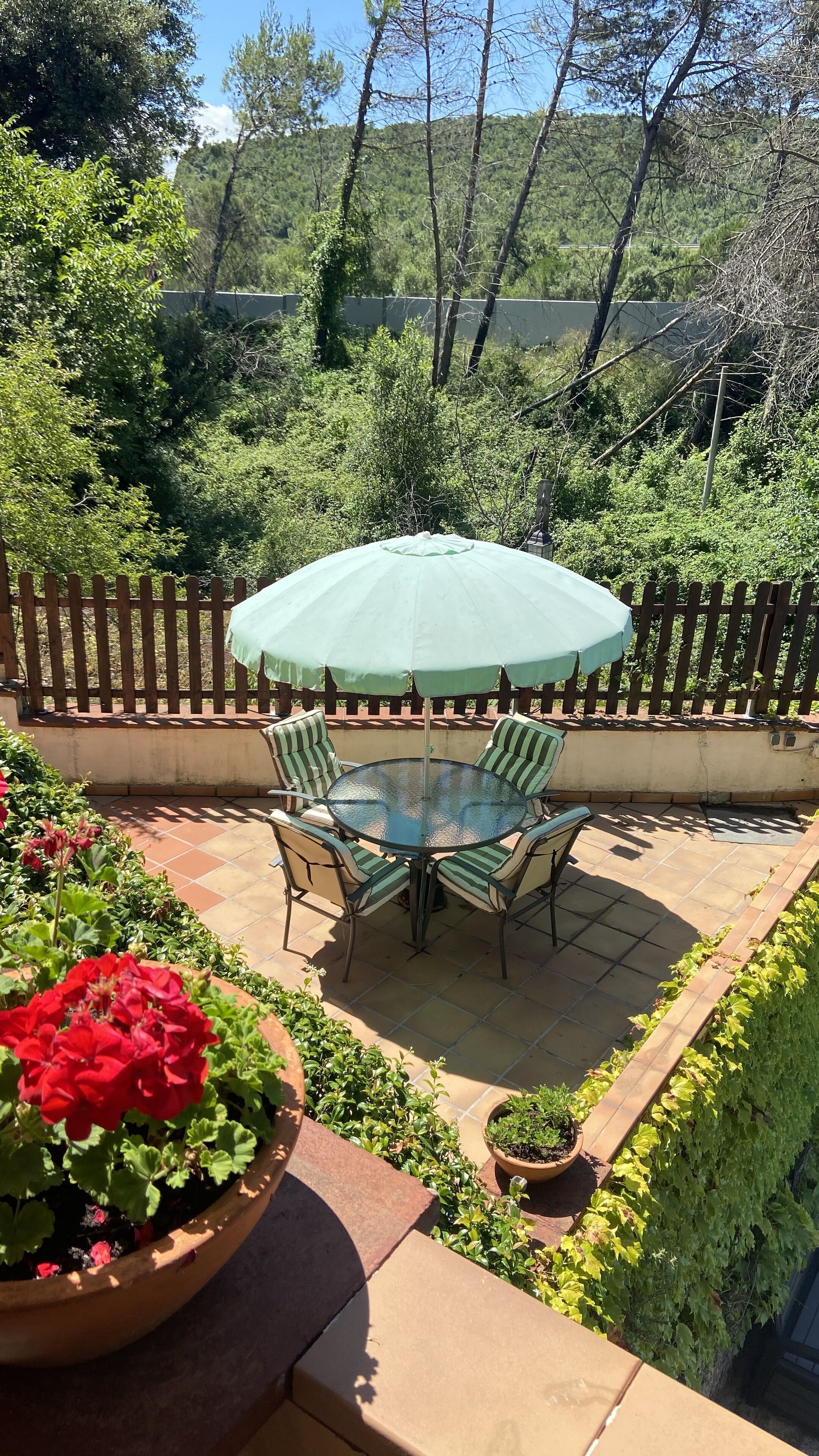 Un patio con un tavolo rotondo in vetro e quattro sedie a righe, un ombrellone e una recinzione in legno.