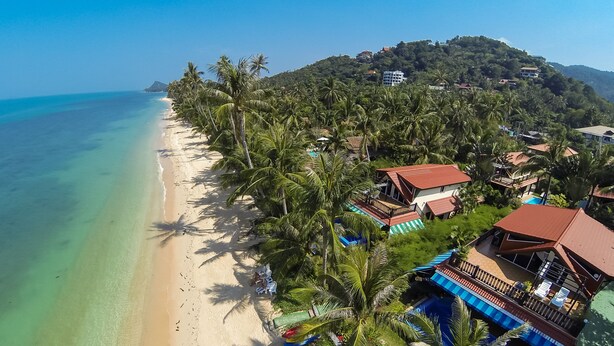 An aerial view of a beach with clear turquoise waters, a sandy shore, and a cluster of buildings with red roofs and balconies overlooking the beach. - Superb Beachfront Villa with private Pool & Jacuzzi in tropical garden. (Ko Samui)