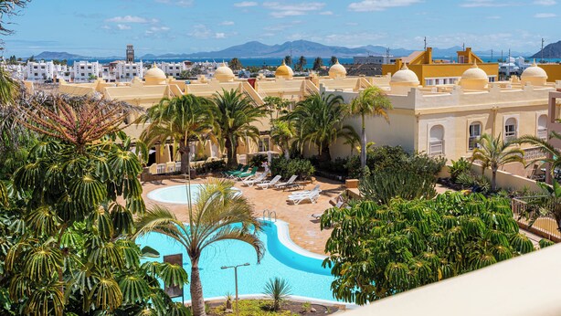 A resort with a swimming pool surrounded by palm trees and lounge chairs, overlooking a coastal cityscape with mountains in the distance. - 3 bedroom villa in sultan complex with wifi/UKTV/pools (Corralejo)