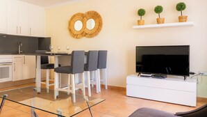 A modern kitchen with white cabinets, a dining area with a glass table and four grey chairs, a flat-screen TV on a white stand, and a wall-mounted shelf with three potted plants. - 3 bedroom villa in sultan complex with wifi/UKTV/pools (Corralejo)