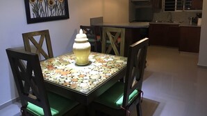 Un comedor con una mesa dispuesta para cuatro personas, rodeada de sillas con cojines verdes. Una vela está colocada sobre la mesa, y un cuadro enmarcado con girasoles cuelga de la pared.