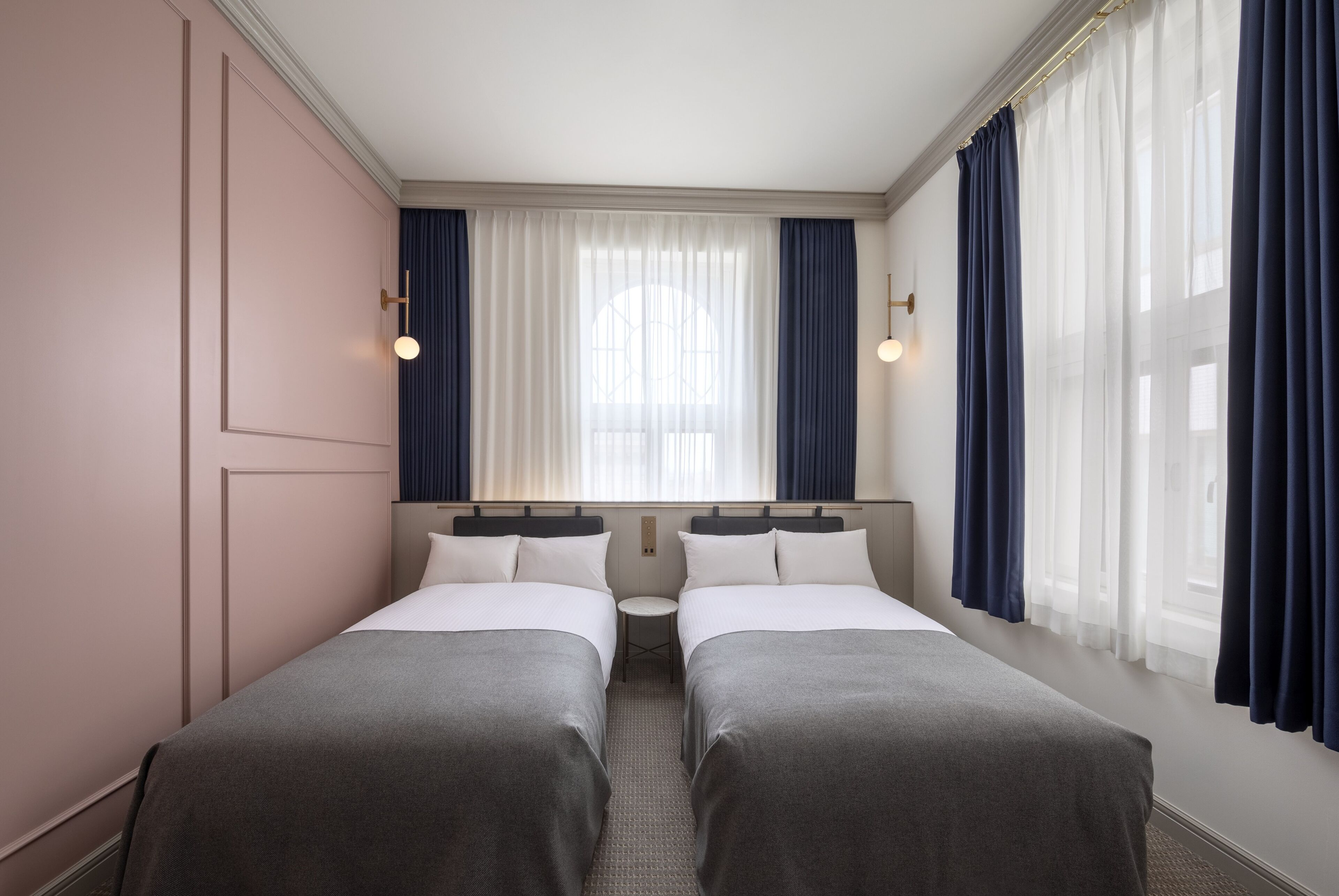 Habitación de hotel con dos camas, una mesita redonda y una ventana con cortinas traslúcidas.