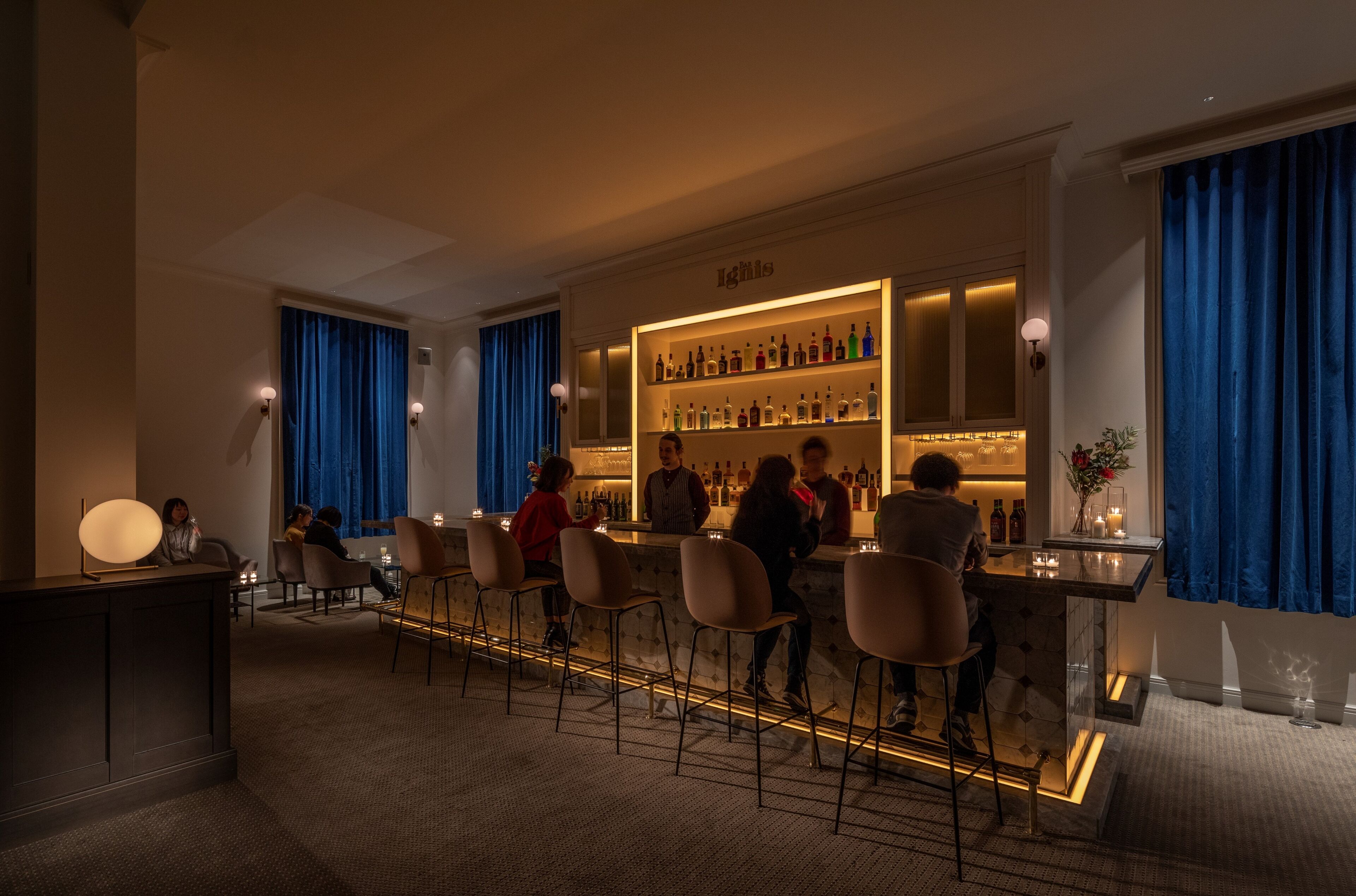 A bar area with a well-lit shelf of bottles, bar stools, and a seating area with a view of the bar.