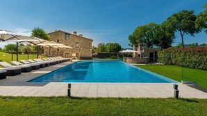 Une piscine rectangulaire, agrémentée de chaises longues et de parasols, entourée d’un bâtiment en pierre et de verdure.