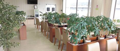 A spacious room with a long table and chairs, surrounded by potted plants, a clock, and a television on the wall.