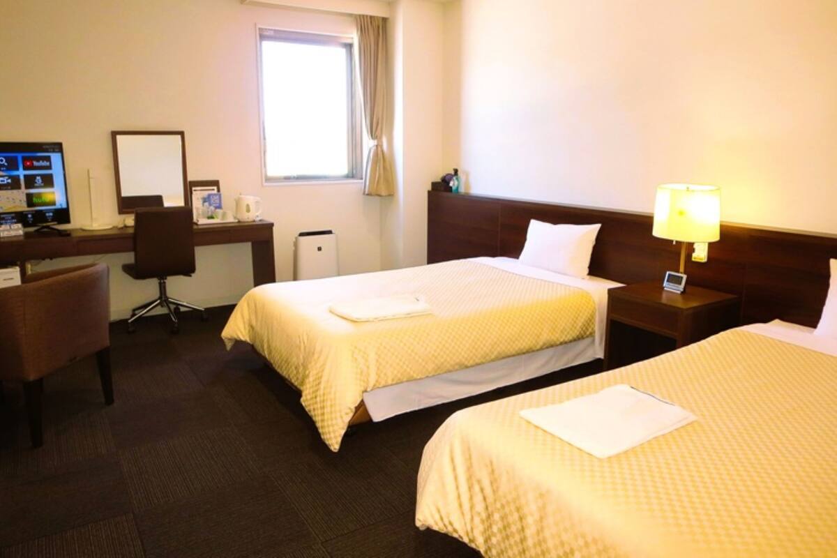 Habitación de hotel con dos camas, un escritorio con computadora, una silla y una ventana con cortinas.