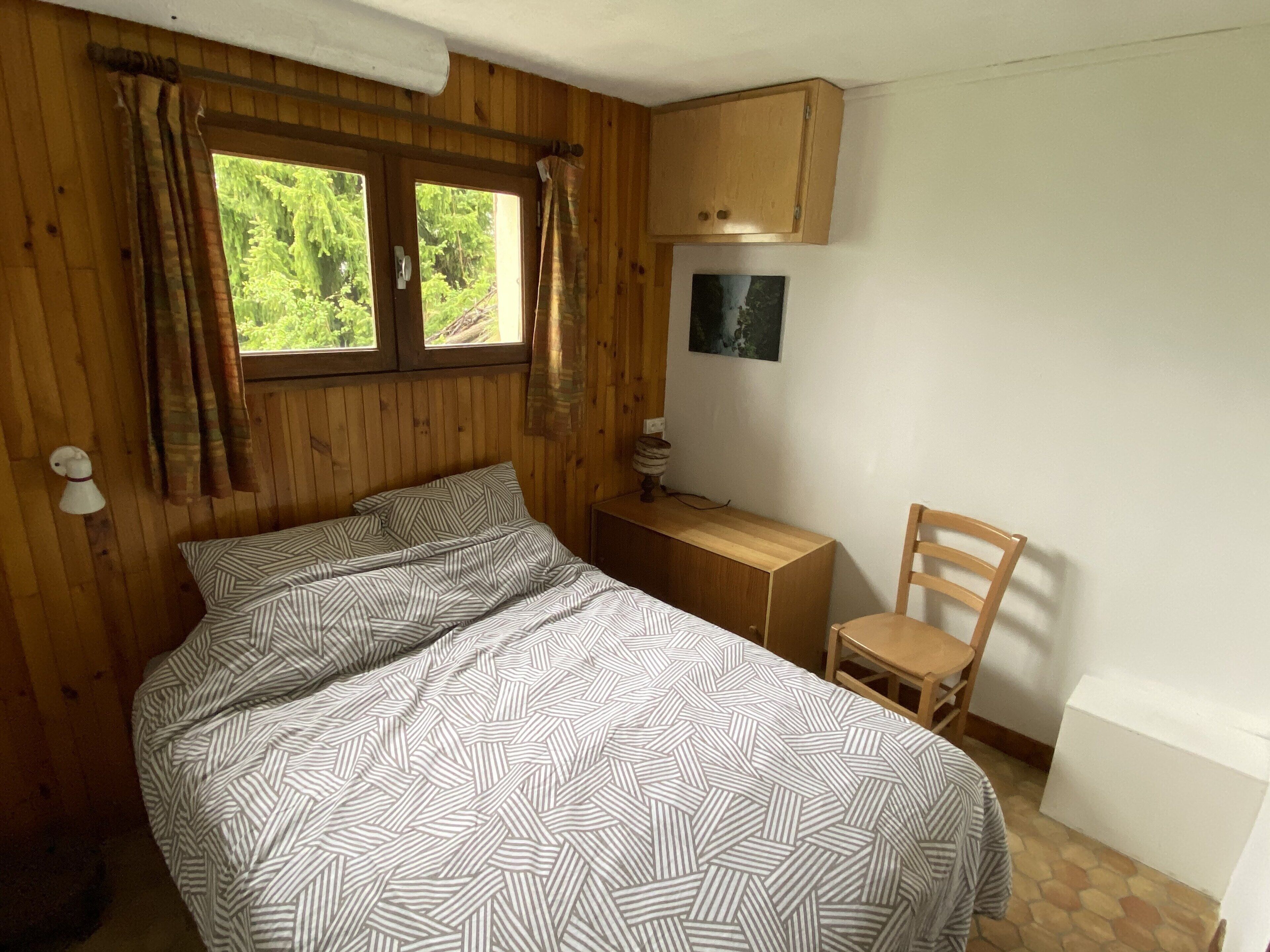 Ein Zimmer mit Holzboden, einem Bett, einem Sessel, einem kleinen Tisch, einem Fenster mit Vorhängen und einem Wandbild.
