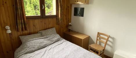 Ein Zimmer mit Holzboden, einem Bett, einem Sessel, einem kleinen Tisch, einem Fenster mit Vorhängen und einem Wandbild.