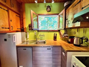 A compact kitchen with a built-in dishwasher, wooden cabinets, a window with a view of greenery, and a clock on the wall.