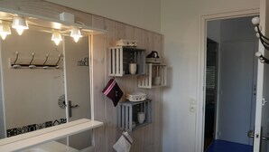 Une salle de bain avec un mur recouvert de panneaux en bois, un lavabo blanc et un miroir avec éclairage intégré. On y trouve des étagères avec des articles de toilette et une serviette accrochée au mur.
