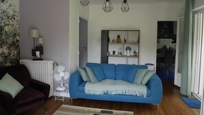 Un salon avec un canapé bleu, un fauteuil rouge, un ventilateur blanc et un radiateur. On y trouve une étagère avec des objets décoratifs et une porte qui donne sur une autre pièce.