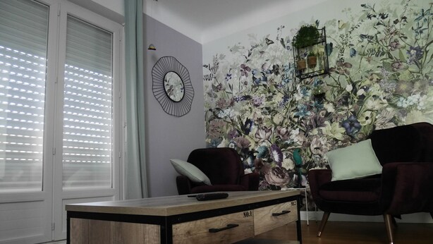 Un salon avec une table basse en bois, deux fauteuils, un miroir fixé au mur et un papier peint à motifs floraux.