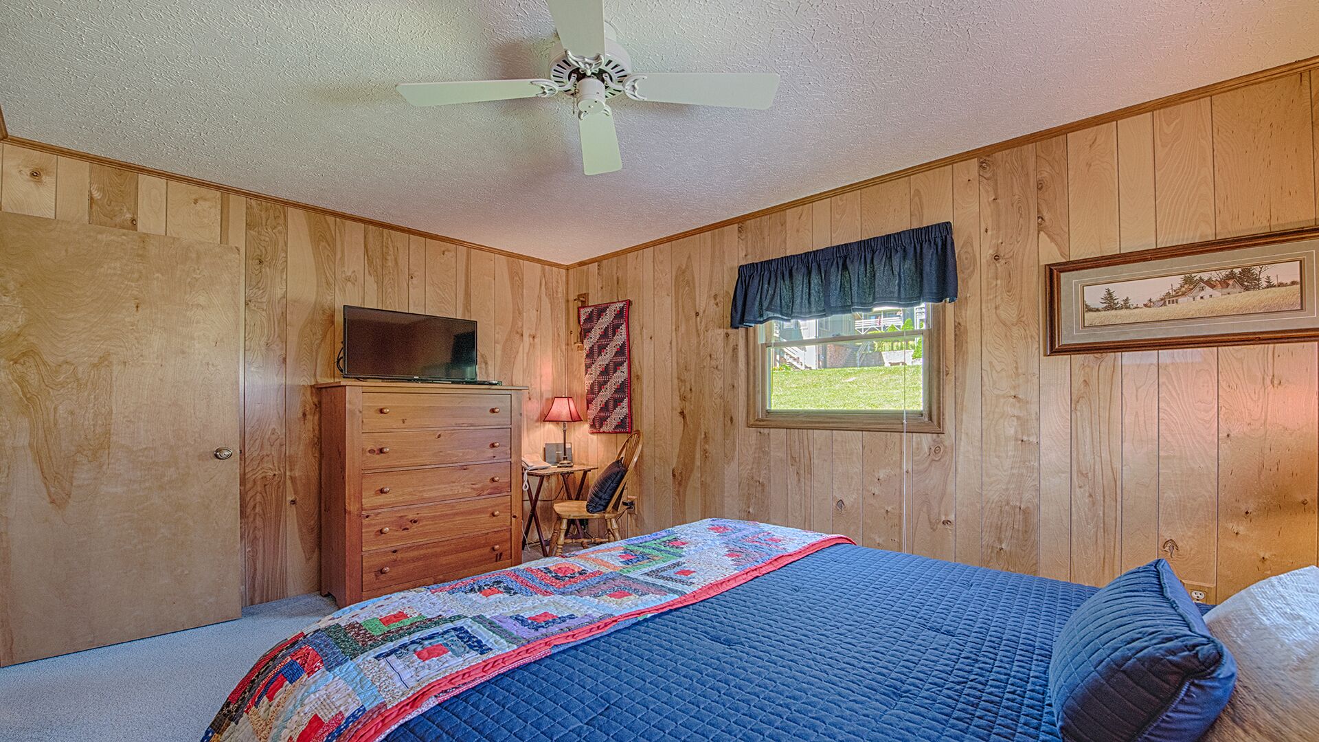 A bedroom with wooden walls, a bed with a quilt, a dresser, a desk with a lamp, a chair, a ceiling fan, and a window with a view.