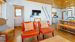 A cozy living room with wooden ceiling, a TV mounted on the wall, and a dining area with a wooden table and benches. - Spacious Home in Town, Back Patio, Shared Pool, Golf Cart Accessible (Port Aransas)