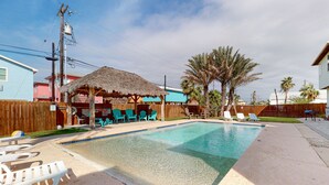 A swimming pool with lounge chairs and a thatched-roof structure. - Spacious Home in Town, Back Patio, Shared Pool, Golf Cart Accessible (Port Aransas)