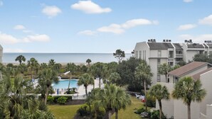 A resort with a swimming pool surrounded by palm trees, a view of the ocean, and multiple-story buildings with balconies.