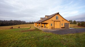 A large wooden barn with a covered porch and a garage, situated in a grassy field with hay bales. - The Mockingbird Farm "Barn-do-minium" is situated on 27 rolling acres. (Seymour)