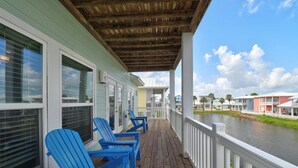 A wooden deck with blue Adirondack chairs, a white railing, and a view of a water body and colorful buildings in the background.
