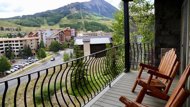 Un balcón con sillas de madera, vista a una montaña y edificios al fondo.