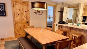 Una mesa de comedor de madera con bancos, una zona de cocina con un lavamanos y encimera, y una puerta de madera con un cartel de bienvenida.