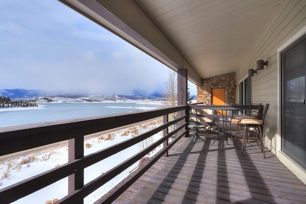 A wooden deck with a stone wall and a table with chairs, overlooking a snowy landscape and a body of water.