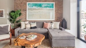A modern living room with a grey sectional sofa, a wooden coffee table, and a large abstract painting on the brick wall. - Modern Surf-Inspired Bungalow with Epic Rooftop Views & Pool (Emiliano Zapata)
