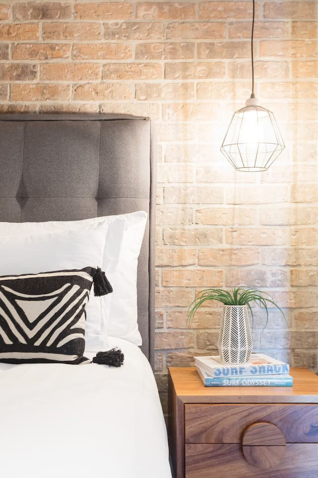 Un lit doté d’une tête de lit rembourrée de couleur grise, de draps blancs et d’un coussin à motifs noir et blanc. Une suspension est fixée au plafond au-dessus du lit, et une petite table de chevet en bois, ornée d’une plante en pot et de quelques livres, se trouve à droite.