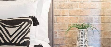 Un lit doté d’une tête de lit rembourrée de couleur grise, de draps blancs et d’un coussin à motifs noir et blanc. Une suspension est fixée au plafond au-dessus du lit, et une petite table de chevet en bois, ornée d’une plante en pot et de quelques livres, se trouve à droite.