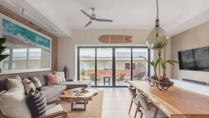 A modern living room with a sofa, coffee table, and dining table. There is a ceiling fan, a large window, and a TV mounted on the wall. - Modern Surf-Inspired Bungalow with Epic Rooftop Views & Pool (Emiliano Zapata)