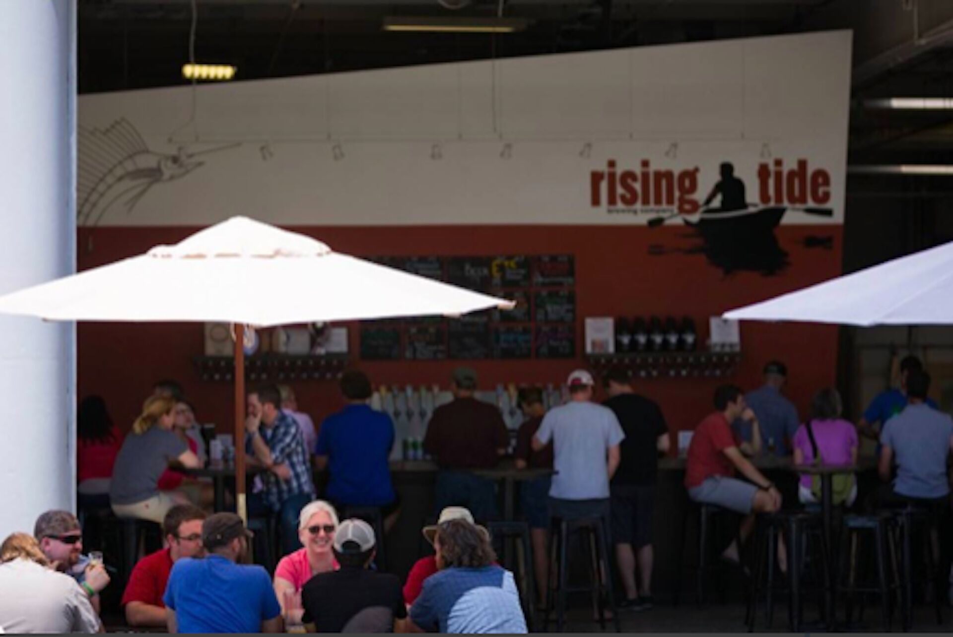 An outdoor seating area with umbrellas, a wall with a 'rising tide' sign, and people dining.